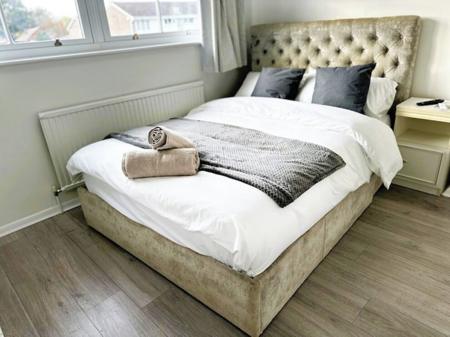 Un lit bien fait, avec une tête de lit beige, une serviette enroulée et une couverture grise.