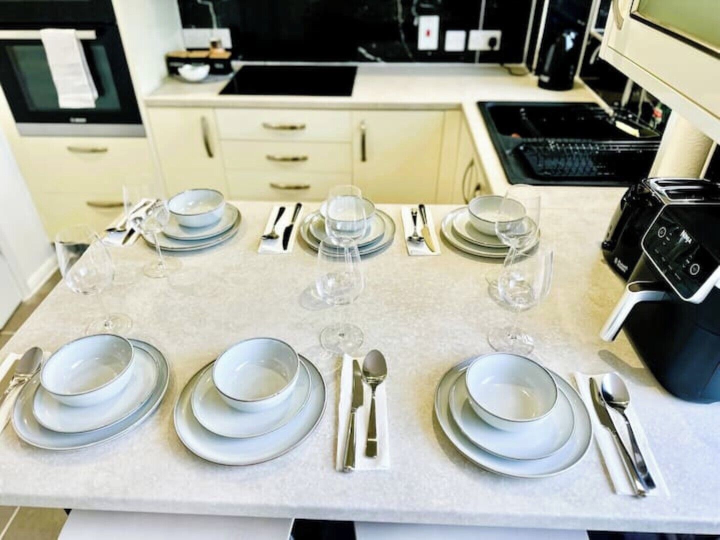A dining table set with plates, glasses, and cutlery in a modern kitchen with a stove and oven in the background.