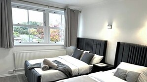 A hotel room with two beds, a window with curtains, a radiator, and a view of trees. - Ama comfort stay (Colchester)