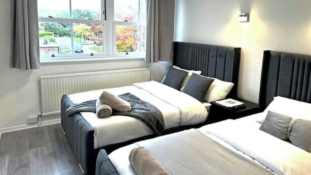 A hotel room with two beds, a window with a view of greenery, a radiator, and a bedside table with a lamp. - Ama comfort stay (Colchester)