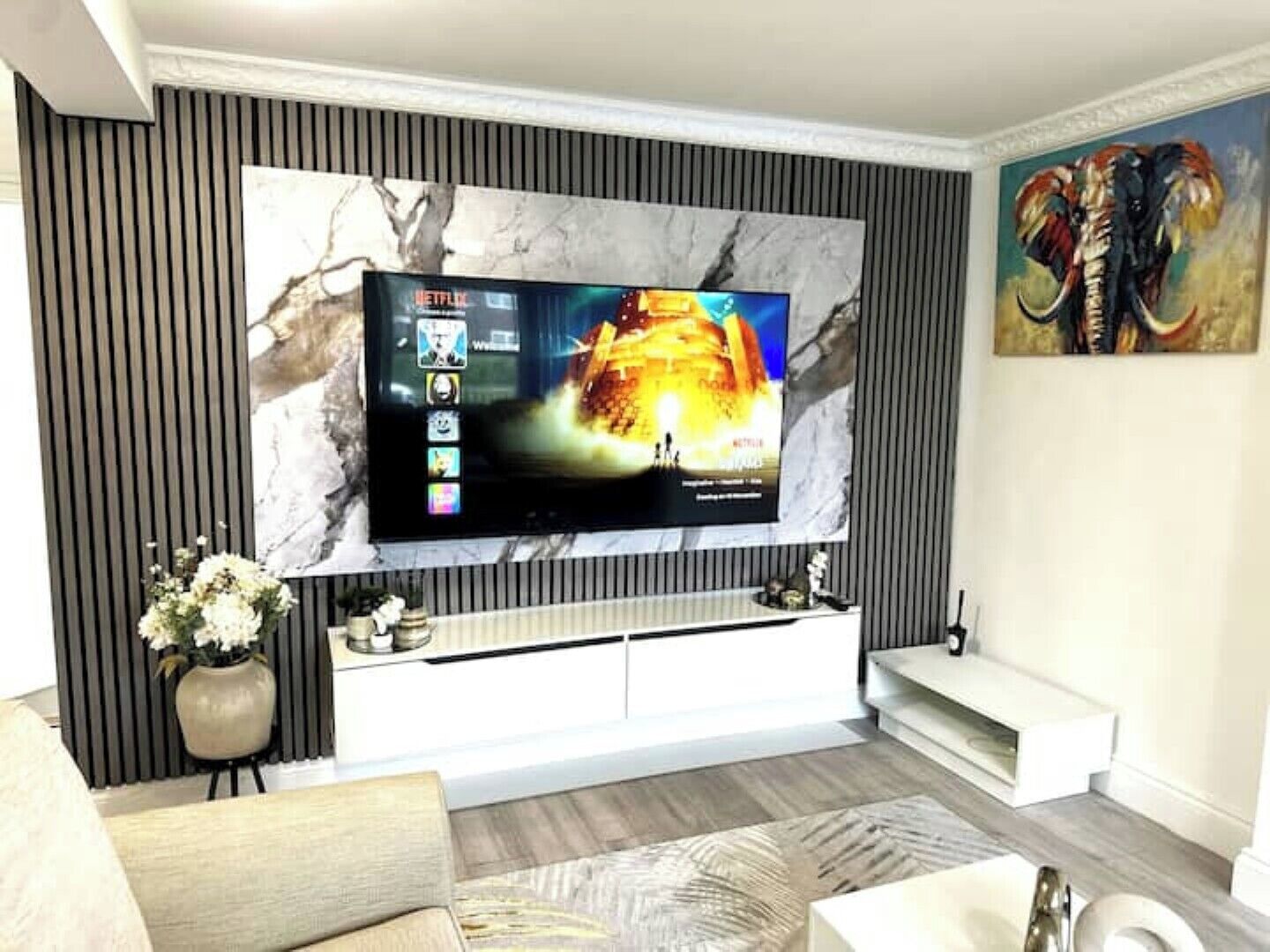 A modern living room with a large flat-screen TV mounted on a marble wall, a white entertainment unit below, and a painting on the right.