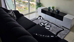 A modern living room with a large window, a black sofa, a white console table, and a flat-screen TV. - Portmore Paradise: Pool & 24/7 Security (Portmore)