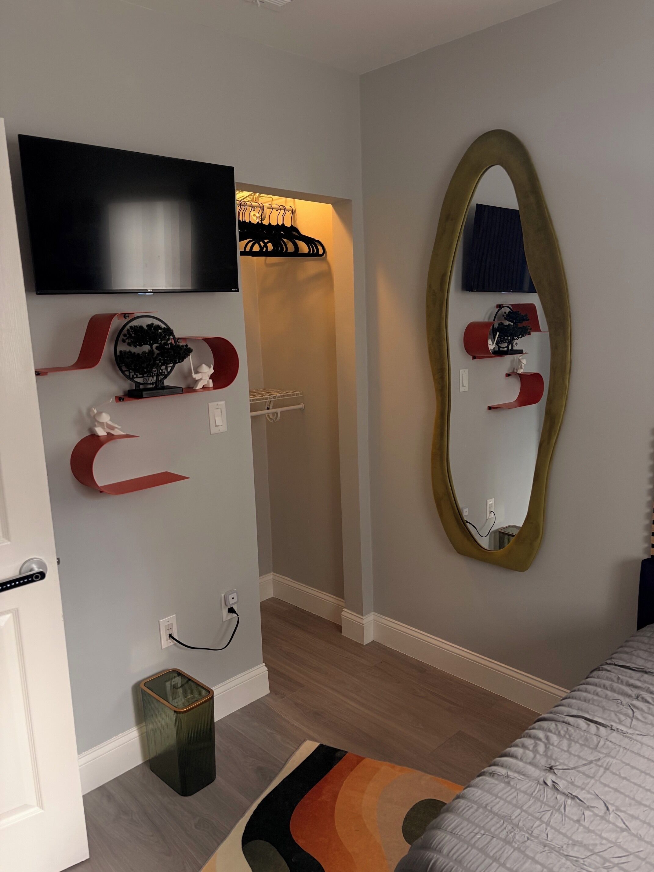 A bedroom with a bed, a TV mounted on the wall, a mirror, and a small trash bin.
