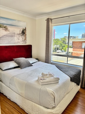 A neatly made bed with striped bedding and a red headboard, a window with a view of buildings and trees, and a painting of a beach scene on the wall. - Immaculate Fully Self Contained Apartment in Central Surfers Paradise (Surfers Paradise)