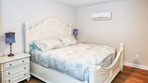 Un dormitorio con una cama blanca, una mesita de noche con cajones, una lámpara y un equipo de aire acondicionado en la pared.