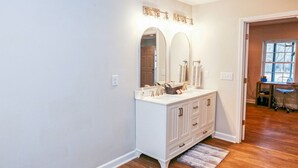 Un baño con armarios blancos, tocador, dos espejos y ventana.