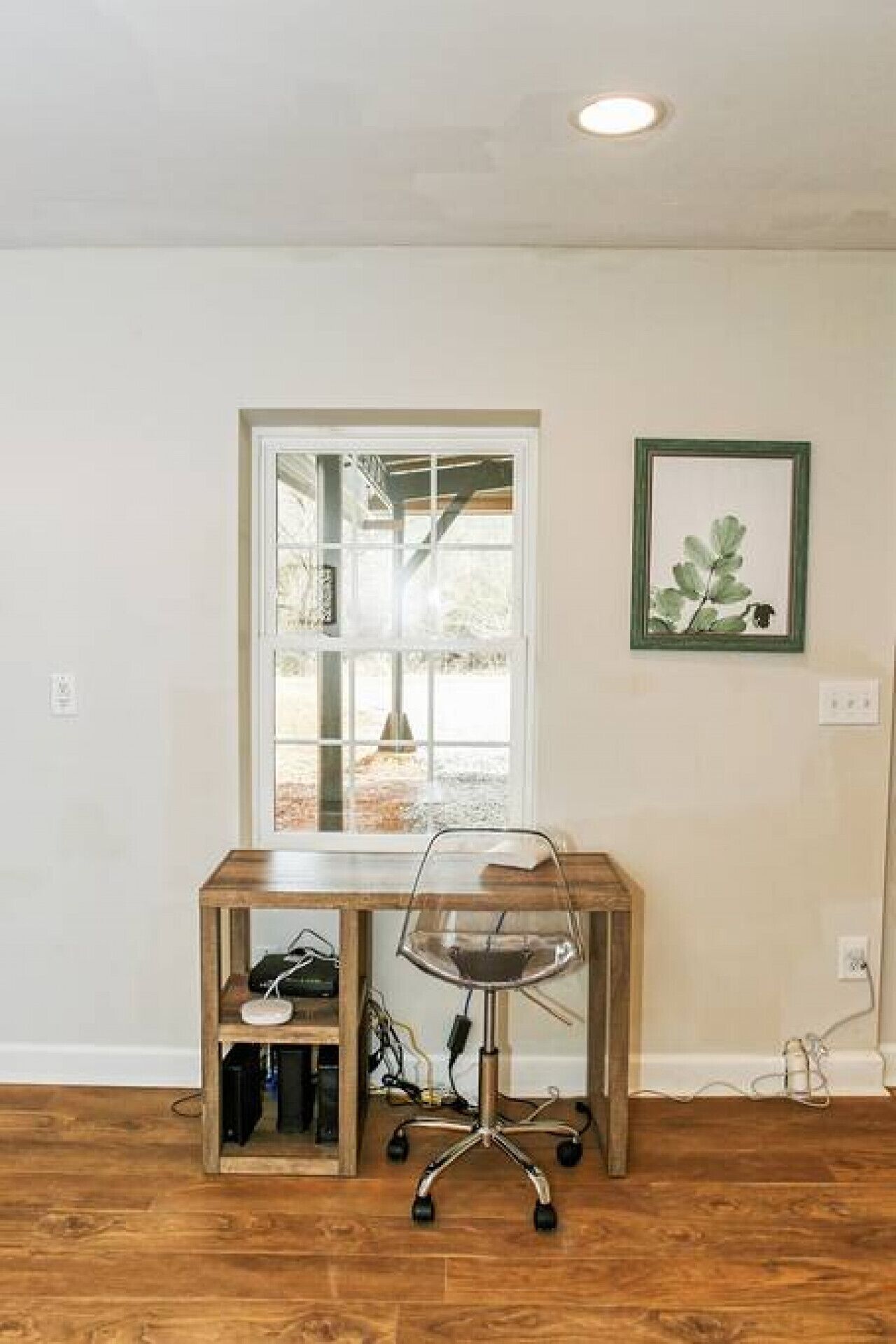 A room with a wooden desk, a chair, a window, and a framed plant.