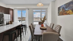 A modern kitchen with a central island, dining area with a long table and chairs, and a large window with a view of a snowy landscape. - Embers Nest - 5 beds, 4 baths, 2 park, Backyard, Thermea Spa (Whitby)