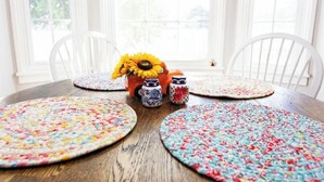 A dining table with four colorful woven placemats, a vase with sunflowers, and two ceramic jars.