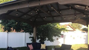 An outdoor patio with a covered seating area, a white picket fence, and a white fence in the background.