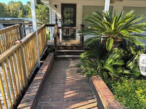 Une terrasse en bois mène à une maison, avec un chemin en briques, un palmier et une pancarte de bienvenue.