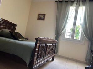 A bedroom with a bed, a nightstand, a window with curtains, and a wall-mounted picture. - Gîte de la Cour - Château Périès (Nissan-lez-Enserune)
