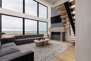 Een moderne woonkamer met een groot raam, een open haard, een trap en uitzicht op het strand.