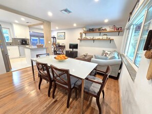 Una cocina moderna con armarios blancos, una mesa de comedor con sillas y una zona de estar con sofá y televisor.