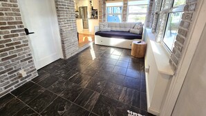 A modern interior with a brick wall, a white door, a black tiled floor, a seating area with a bench and cushions, and a small wooden stool. - Peaceful 3 Bedroom 2 Bathroom Pool Home (Hollywood)