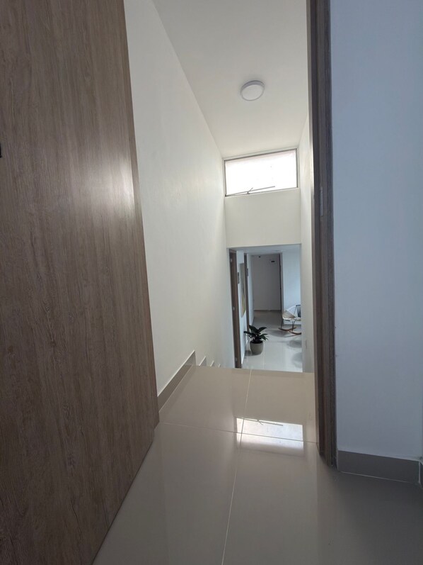 A hallway with a wooden floor, white walls, and a ceiling light. There is a potted plant and a chair visible in the distance. - Hotel Portal Del Cerro (Manizales)