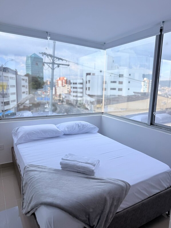 Habitación de hotel moderna con una cama grande, ropa de cama blanca y vistas de la ciudad a través de amplias ventanas.