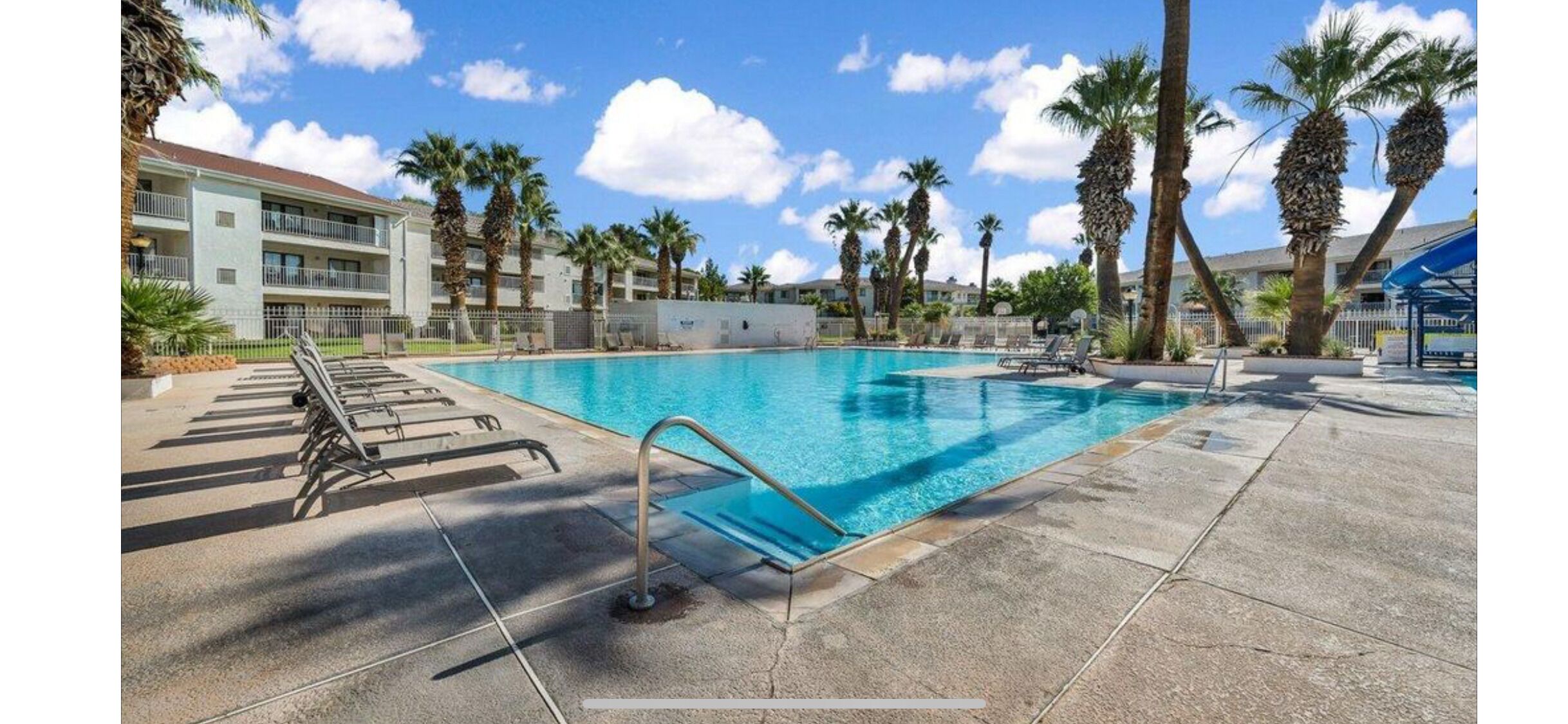 A swimming pool with lounge chairs, palm trees, and a building in the background.