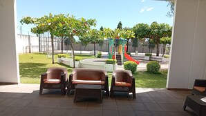 Un patio con tres sillas de mimbre y una pequeña mesa, con vista a un parque infantil que tiene un tobogán y estructuras para escalar.