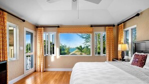 Una camera da letto con un letto grande, vista su palme e oceano, e una ventola a soffitto.
