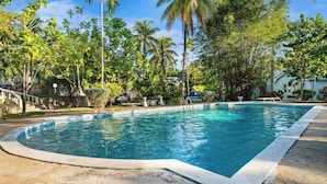Una piscina rettangolare circondata da vegetazione lussureggiante e palme, con un vialetto in pietra che conduce a un edificio bianco.