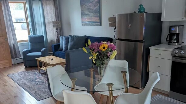 A modern living room with a glass dining table, white chairs, a sofa, and a kitchen area with a refrigerator and coffee maker. - Entire House. 2+ bed, 2 bath near Lake & bikeway (Burlington)