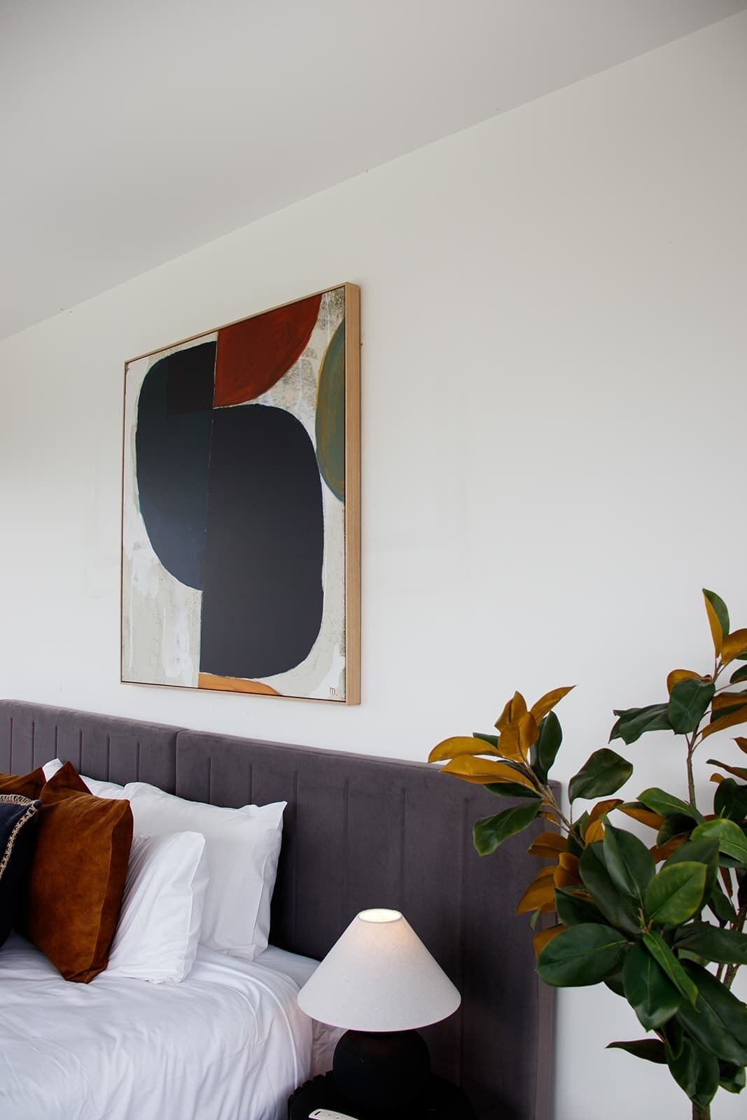 A bedroom with a grey upholstered headboard, a white bed with multiple pillows, a bedside lamp, and a large abstract painting on the wall.