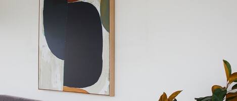 A bedroom with a grey upholstered headboard, a white bed with multiple pillows, a bedside lamp, and a large abstract painting on the wall.