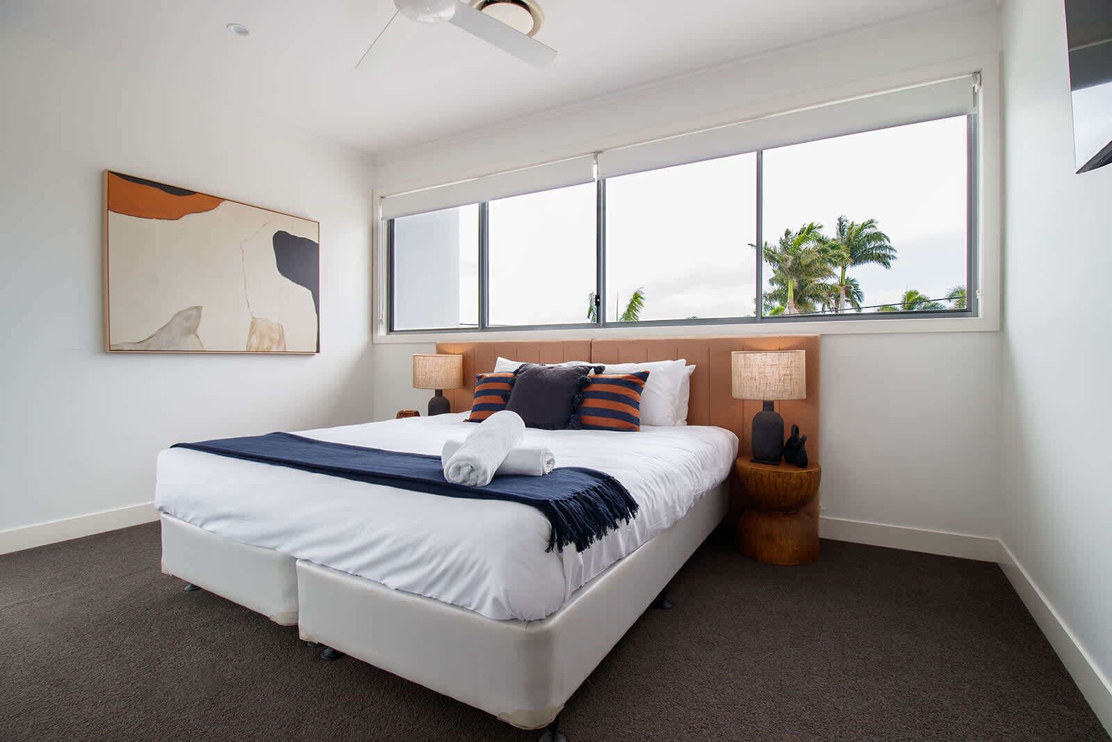 A modern hotel room with a large bed, a view of palm trees, and a ceiling fan.