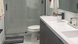 Un baño moderno con cabina de ducha de cristal, inodoro blanco, espejo grande y lavamanos blanco con muebles de madera oscura.
