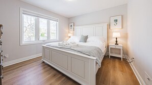 A bedroom with a large bed, a window, a bedside table, and a wall-mounted picture. - Spacious, Newly Renovated, Centrally Located | TMH (Burlington)