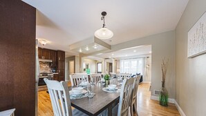 A spacious dining area with a wooden table set for six, surrounded by white chairs. A kitchen with dark wooden cabinets and a stainless steel range is visible in the background. - Spacious, Newly Renovated, Centrally Located | TMH (Burlington)
