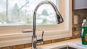 A kitchen with a stainless steel faucet, a window with a view of trees, and a bottle of dish soap on the counter. - Spacious, Newly Renovated, Centrally Located | TMH (Burlington)