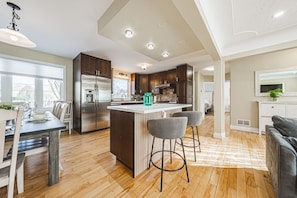 A modern kitchen with a central island, bar stools, and a dining area with a table and chairs. - Spacious, Newly Renovated, Centrally Located | TMH (Burlington)