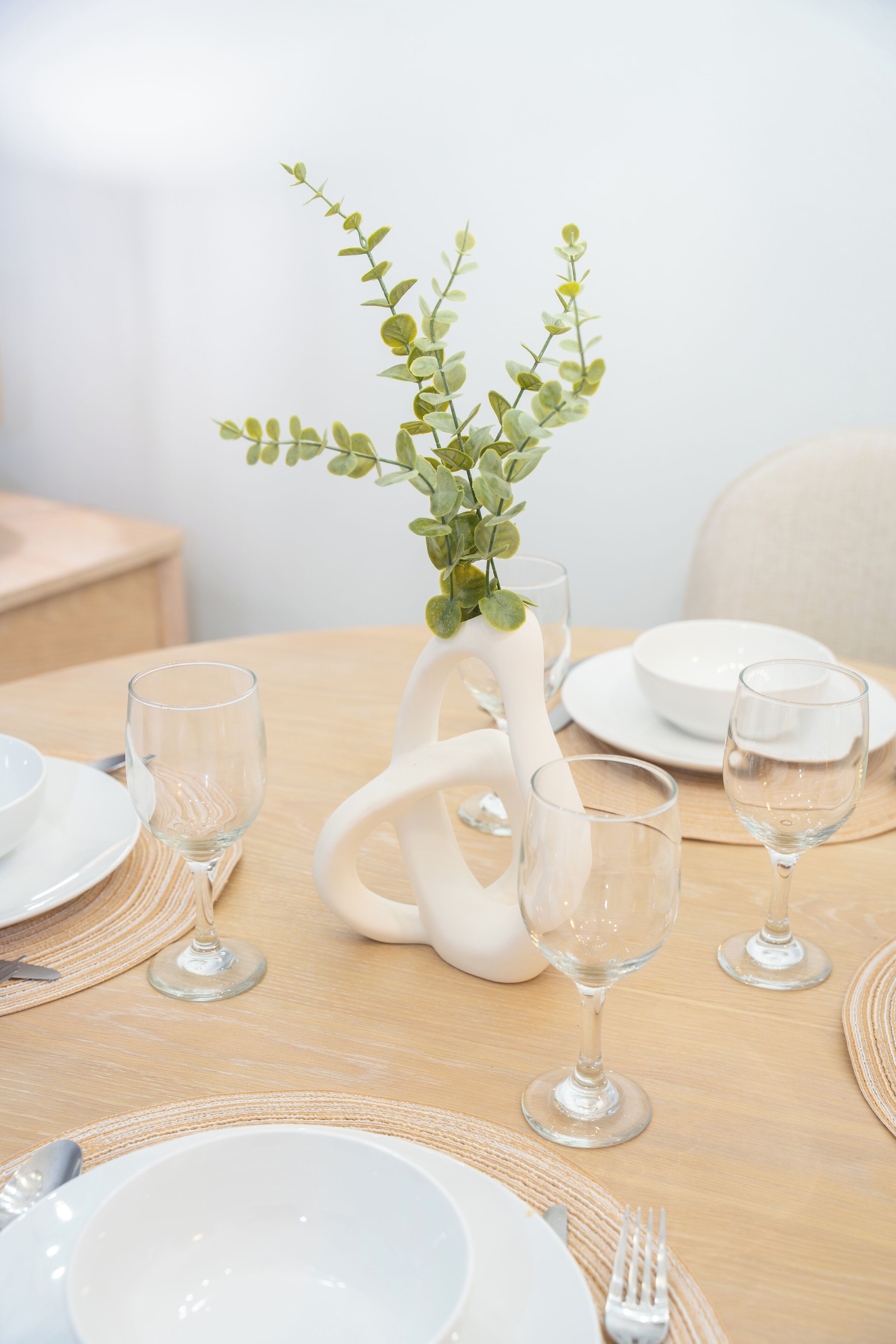 Een tafel gedekt met witte borden, kommen en wijnglazen, met een witte vaas met groene bladeren.