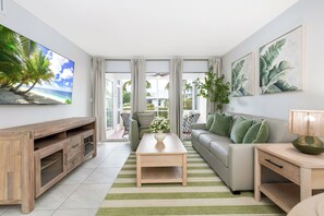 Een moderne woonkamer met een houten tv-meubel, een bank met groene kussens, een salontafel en een televisie waarop een strandscène te zien is.
