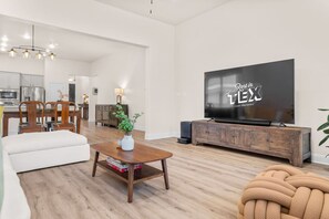 A modern living room with a flat-screen TV, wooden furniture, and a dining area in the background. - Spacious 3BR Getaway w Dedicated Office | Sleeps 5 (Leander)