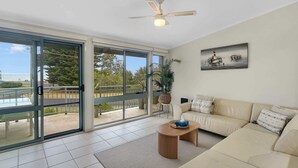 A spacious living room with a beige sofa, a coffee table, and a ceiling fan. Large sliding glass doors lead to an outdoor balcony with a view of the sea and trees.