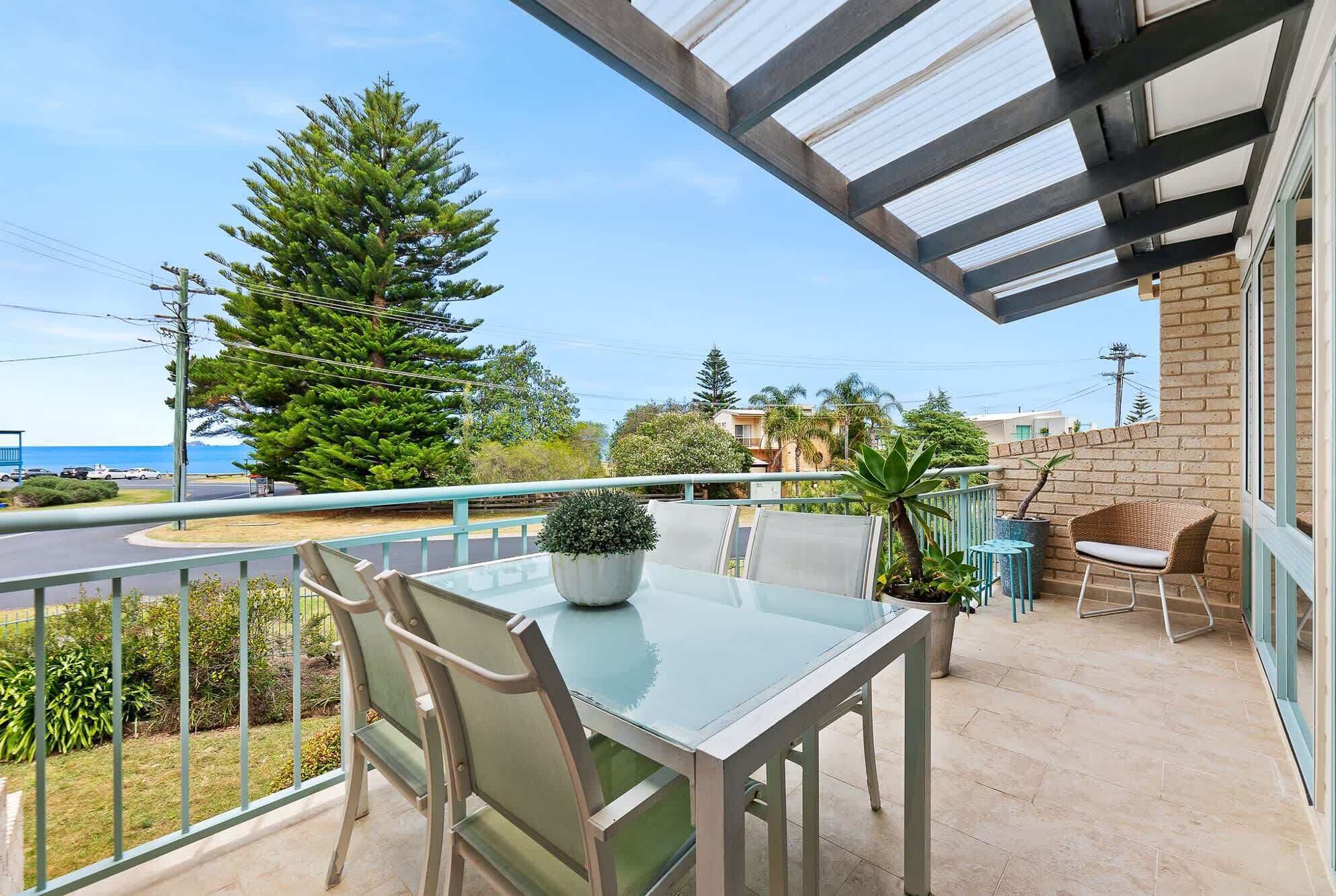 A balcony with a glass table and chairs, potted plants, and a view of the street and trees.