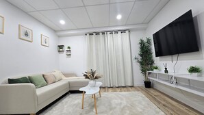 Una sala de estar moderna con un sofá, una mesita de centro blanca, un televisor de pantalla plana en la pared, un ventanal con cortinas y una planta en maceta.