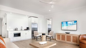 Eine moderne Küche mit weißen Schränken, einem eingebauten Backofen und einem Kühlschrank. Ein runder Esstisch mit zwei Stühlen, ein Flachbildfernseher an der Wand und ein Weidenkonsolentisch mit einer Schüssel.