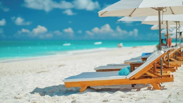 Ein Strand mit weißem Sand und klarem, türkisblauem Wasser. Holzliegen mit weißen Sonnenschirmen sind in Reih und Glied aufgereiht.