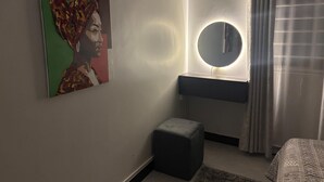 A bedroom with a bed, a bedside table, a round mirror with a light, a small ottoman, and a painting on the wall. - Sky View 1-bedroom apartment in the heart of Kampala (Kampala)
