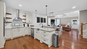 A spacious kitchen with white cabinetry, a central island, and a dining area with a brown leather couch. - Conroe 4BR near The Woodlands | Arcade Game Room, Gym, Playground, Sleeps 13 (Conroe)