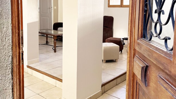 An interior view of a room with a wooden door, a tiled floor, a brown armchair, a white ottoman, and a glass table with chairs.