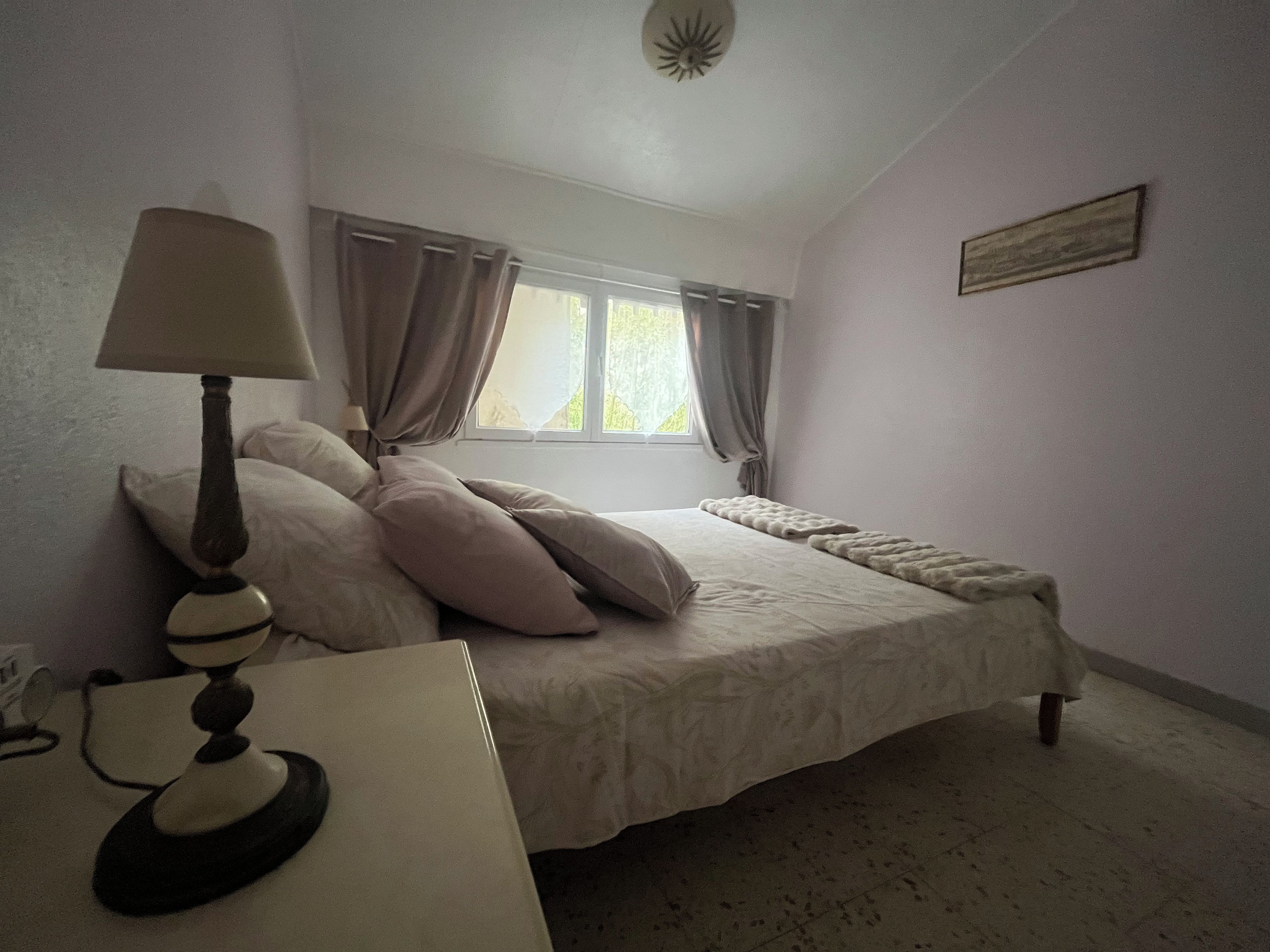 A bedroom with a bed, a bedside lamp, a window with curtains, and a wall-mounted picture.