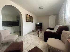 Una sala de estar con televisor, sofá y sillón, una cocina al fondo y un cuadro en la pared.
