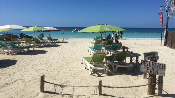 A beach with rows of green and white lounge chairs under umbrellas, a sign reading "Fancy People Best Beach Ever", and a wooden fence in the foreground. - Garden-Sea view, 2 bedroom/2bath Montego Freeport (Montego Bay)