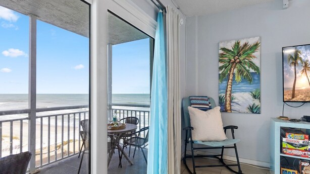 A balcony with a view of the beach, a table with chairs, a rocking chair, and a cabinet with board games. - Oceanfront 2BR Condo with Large Balcony & Beach Access! 🌴 (Satellite Beach)