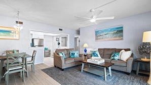 A modern living room with a ceiling fan, a dining area with a table and chairs, a kitchen with a refrigerator, and a beach-themed painting on the wall. - Oceanfront 2BR Condo with Large Balcony & Beach Access! 🌴 (Satellite Beach)